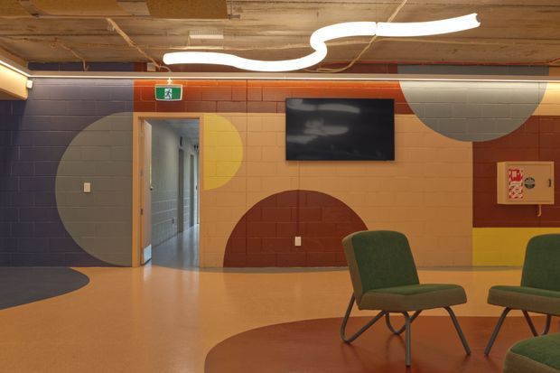 A colourful contemporary commercial interior with a tiled feature wall painted in large overlapping geometric blocks of navy, orange, mustard yellow, cream, and terracotta. The space has an exposed unfinished ceiling with visible services, sculptural white ceiling lighting, a wall-mounted television, an open doorway, and green upholstered lounge chairs on a smooth warm-toned floor.