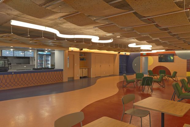 A spacious, vividly designed commercial interior resembling a school or institutional cafeteria and common area. The room features a service counter with digital menu screens and refrigerated display units, extensive built-in timber cabinetry, colourful geometric wall graphics, a patterned counter front, scattered green chairs and timber tables, and a sculptural suspended ceiling with exposed acoustic panels and flowing linear lighting.