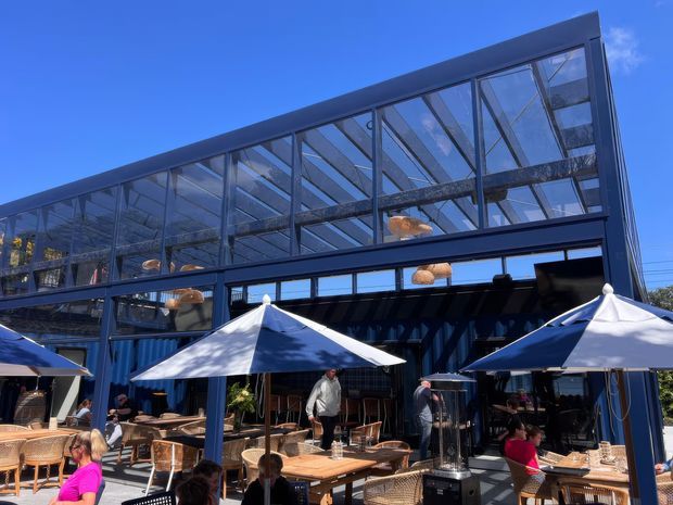 Exterior view of a contemporary multi-level commercial venue with a dark steel-framed glass upper storey and open glazed frontage. The ground-level outdoor dining terrace has white shade umbrellas, timber tables, woven rattan chairs, and patrons seated beneath a clear blue sky.