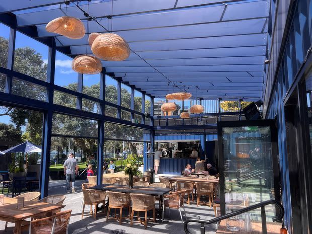 Bright contemporary hospitality venue with extensive floor-to-ceiling glazing, a translucent roof canopy, dark structural framing, and an open café or restaurant dining area. Woven pendant lights hang above timber tables and woven chairs, with a bar and mezzanine-style upper area at the rear. Outdoor greenery and seating are visible beyond the glazing.