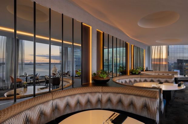 A sophisticated high-rise dining or lounge interior with sweeping floor-to-ceiling glazing overlooking a waterfront cityscape. Curved banquette seating in patterned upholstery wraps around round tables, while dark slender window mullions, warm concealed lighting, sculptural recessed ceiling details, indoor plants, and panoramic views create an upscale hospitality atmosphere.