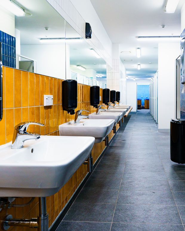 A bright commercial bathroom or washroom with a long row of white wall-mounted basins and chrome taps along a mustard-orange tiled wall. Large mirrors run above the basins, with black wall-mounted soap or hand-dryer dispensers. The space has dark grey tiled flooring, white tiled walls, fluorescent lighting, and toilet cubicles extending along the right side.