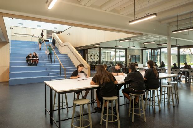 A bright contemporary school or educational commons with students studying at long communal tables and others seated on a broad blue stepped stair. The open-plan space features exposed ceiling services, suspended linear lights, extensive interior glazing, pale timber framing, dark flooring, and a stair connecting different levels.