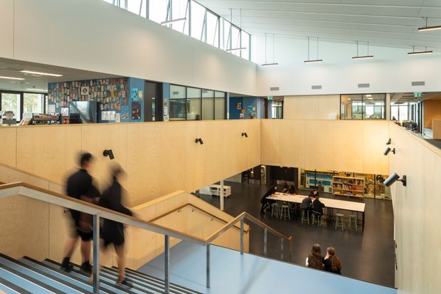 A bright, contemporary multi-level institutional interior viewed from a stair landing. Pale timber-clad balustrades and partitions frame an open lower-level area with tables, chairs, shelving and visible work or study spaces. Large internal glazed partitions, clerestory glazing, linear pendant lights and a high white paneled ceiling create an airy atrium-like environment. People are moving on the stairs and seated below.