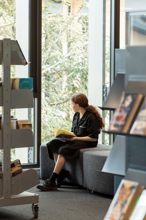 A bright contemporary library or reading area with full-height glazing overlooking leafy vegetation. A woman in dark clothing sits reading on a large grey upholstered built-in or modular bench, while angled book display shelving frames both sides of the image. The space has a calm, airy atmosphere with abundant natural light and muted neutral finishes.