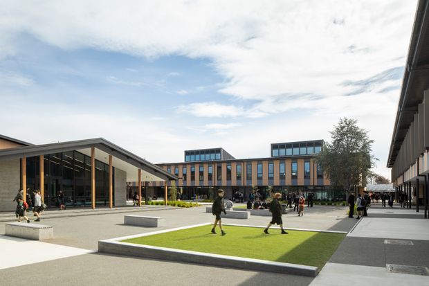 A contemporary institutional courtyard or campus plaza viewed from outdoors. The open paved space includes raised rectangular lawn areas, concrete seating blocks, and groups of people, surrounded by multi-storey buildings with extensive vertical glazing, dark metal framing, and flat roof forms. A covered entrance pavilion with tall glazed walls occupies the left side, while the central building has rooftop clerestory-like volumes.
