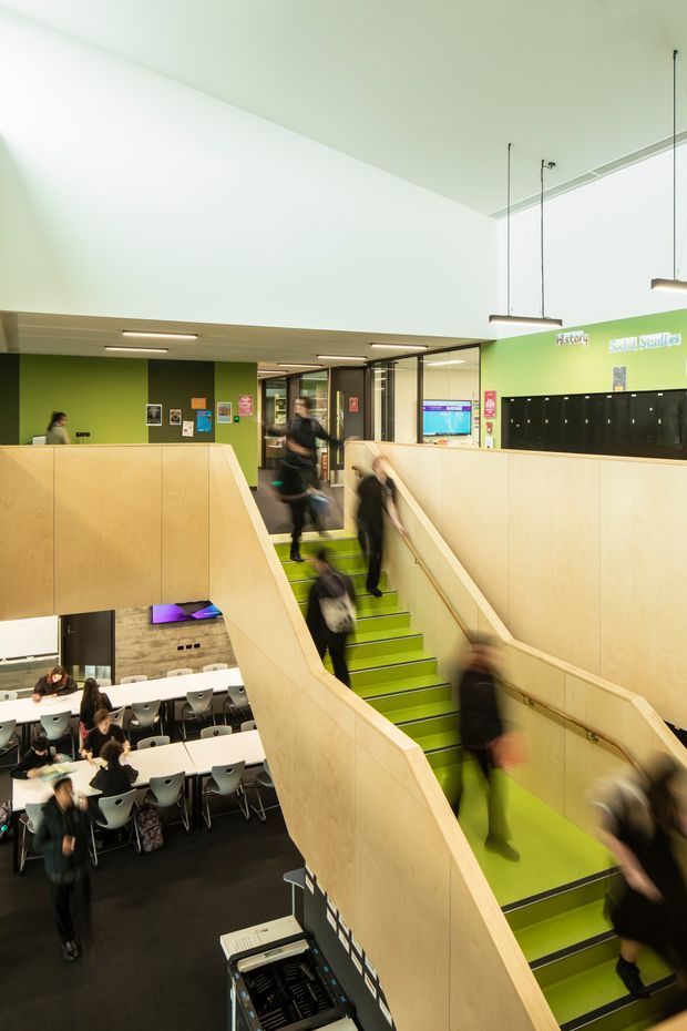 A bright, contemporary multi-level educational or commercial interior with a prominent lime-green stairway and ramp enclosed by pale timber balustrade walls. The lower level contains communal dining or breakout tables and chairs, while the upper landing has black lockers, graphic wall signage, display screens and glazed partitions. Suspended linear pendant lights and clean white ceilings create an airy, functional atmosphere. Motion-blurred people occupy the stairs and lower seating area.