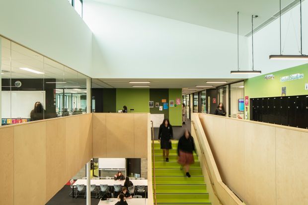 Interior view of a contemporary multi-level institutional or educational space, centred on a broad lime-green staircase with timber-clad balustrades and partitions. Glazed walls and mirrors open views into adjacent rooms, including a lower-level dining or common area. The space has high ceilings, suspended linear lighting, bright green accent walls, and black locker storage.