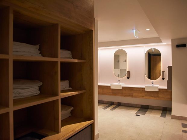 A warmly lit bathroom vanity area viewed past open built-in timber shelving stocked with folded towels. The bathroom features two arched mirrors, wall-mounted white basins, timber vanity cabinetry, pale tiled flooring, and dark rectangular accent tiles arranged along the floor perimeter.