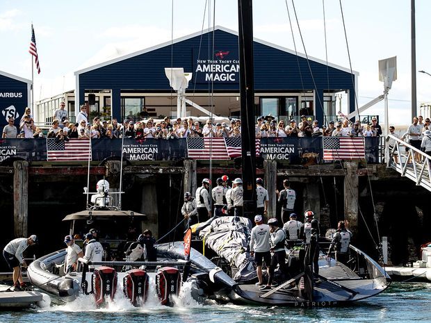A busy waterfront marina scene centered on a dark blue-clad yacht club or race-team building with a gabled roof and large glazed openings. Spectators gather on the pier and balconies above a racing powerboat being serviced at the dock, with nautical rigging and American-themed event branding visible throughout.
