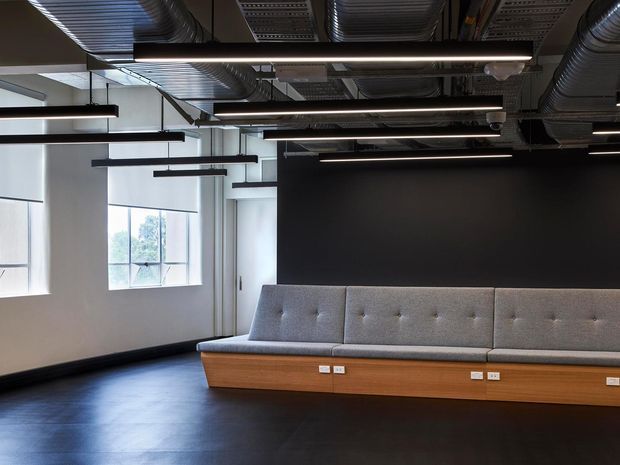 A modern commercial interior with an exposed services ceiling, suspended linear LED lighting, black feature wall, dark flooring, and a long built-in upholstered bench with timber base and integrated power outlets. Large windows provide natural light.