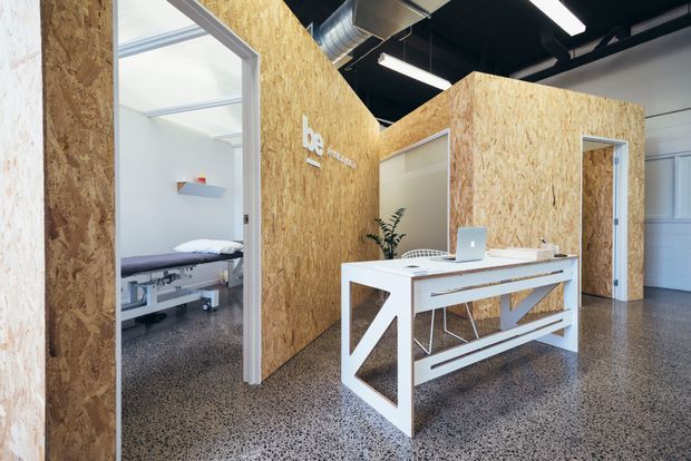 Contemporary commercial reception or office interior with a white reception desk set against exposed oriented strand board partition walls. The space has polished concrete or terrazzo-like flooring, an exposed black ceiling with ductwork and linear fluorescent lighting, and a treatment or examination room visible through an open doorway.