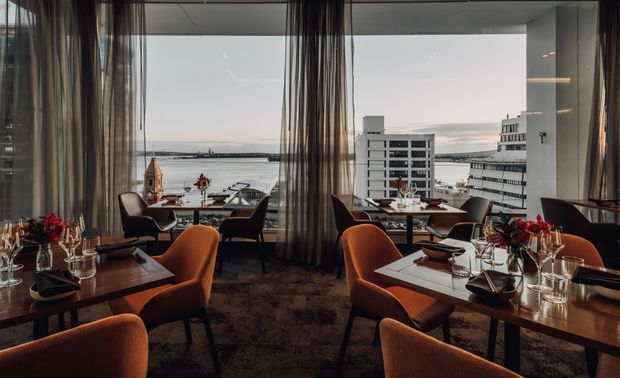 A refined restaurant dining room with floor-to-ceiling windows overlooking a waterfront harbour and city buildings. Dark timber dining tables are set with glassware and red floral arrangements, while rust-orange upholstered chairs, sheer drapery, and warm ambient lighting create an elegant atmosphere.