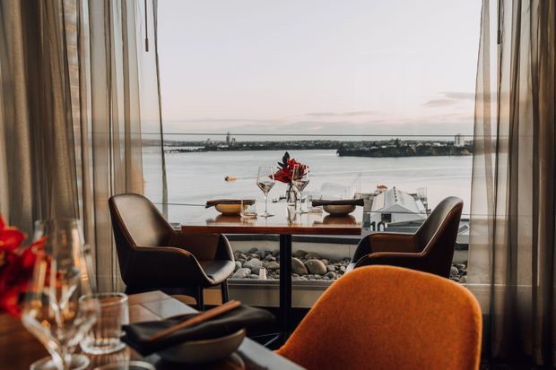 A refined dining area beside full-height glazing, with a small dark dining table set for two, sculptural dark armchairs, and a prominent burnt-orange upholstered chair in the foreground. Sheer curtains frame expansive waterfront views, creating an intimate yet luxurious restaurant-like atmosphere. Natural stones line the lower window edge, with floral arrangements and tableware adding decorative detail.