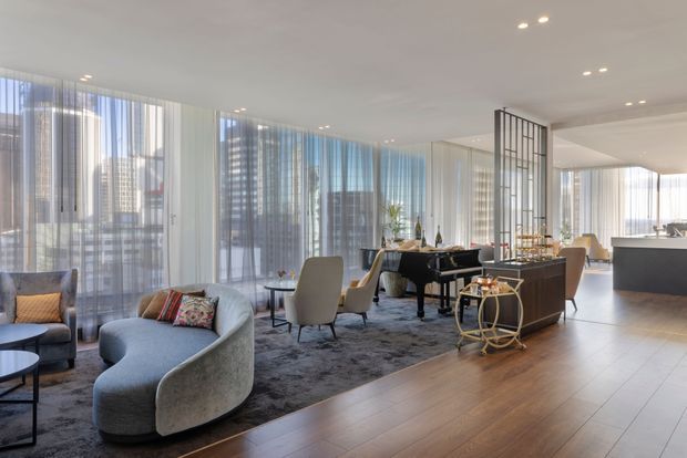 A bright, open-plan contemporary apartment interior featuring a curved grey sofa, lounge chairs, dark dining table, and a large rug over warm timber flooring. Floor-to-ceiling sheer curtains provide extensive glazing and city views, while a black-framed glazed screen subtly separates the dining and living areas from the kitchen beyond.