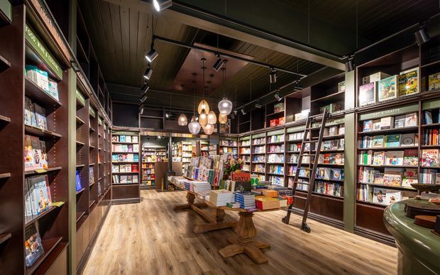 A warmly lit bookstore interior with floor-to-ceiling dark timber shelving filled with books and magazines, a central wooden display table, decorative pendant lights, track lighting, and a wood-look floor. A rolling library ladder leans against the shelving on the right.