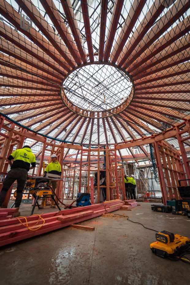 Interior construction view of a large circular or polygonal timber-framed structure under construction. Radiating reddish timber roof members converge around a prominent central circular skylight or roof opening, with extensive temporary scaffolding and safety mesh. Construction workers in high-visibility clothing, tools, timber framing, and concrete flooring are visible below.