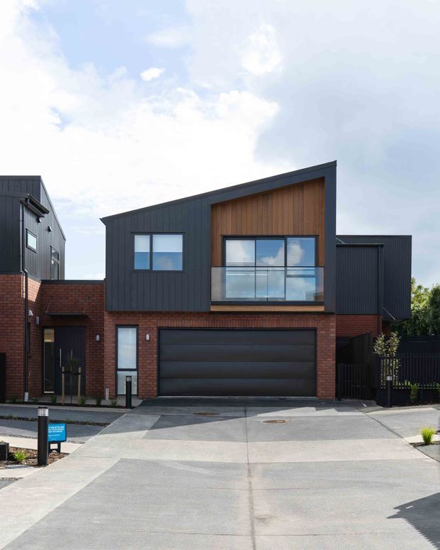 Street-facing view of a contemporary multi-storey townhouse with a dark vertically clad upper facade, warm timber-lined gable feature, red brick lower level, large glazed windows, integrated double garage, concrete driveway and dark fencing.
