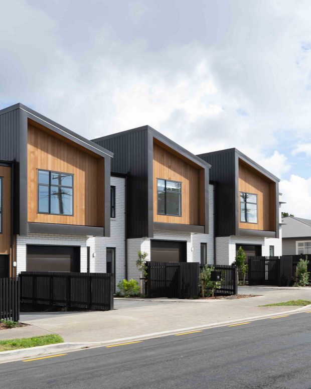 Street-facing view of a row of contemporary multi-storey townhouses. The buildings feature dark vertical cladding, warm timber-look recessed upper façades, white brick lower façades, attached garages, black fencing, and pitched roof forms.