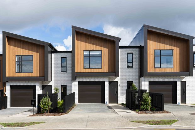 Street-facing view of a row of contemporary multi-storey townhouses. The buildings feature white brick facades, dark framed garage doors and windows, and prominent upper-level projecting boxes clad in warm vertical timber. Each home has a narrow driveway, small landscaped frontage, and a strongly geometric facade beneath a pitched or angled roof form.