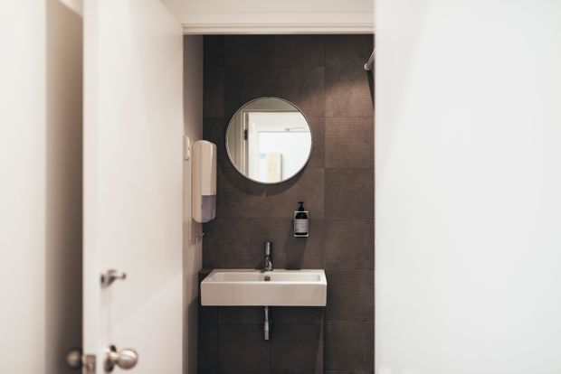 Compact bathroom viewed through a partially open door, featuring a wall-mounted white basin, round mirror, dark textured wall tiles, and minimal fixtures. The space has a clean, contemporary, slightly moody appearance.