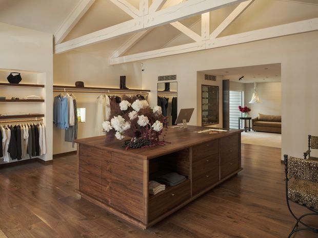 A warm, refined walk-in wardrobe with extensive open clothing racks and shelving along the walls, centered around a large dark timber island with drawers and open compartments. White-painted exposed roof trusses and beams create a high ceiling, while a doorway opens to an adjacent lounge area. The room features polished timber flooring, neutral walls, a large mirror, and patterned accent seating.