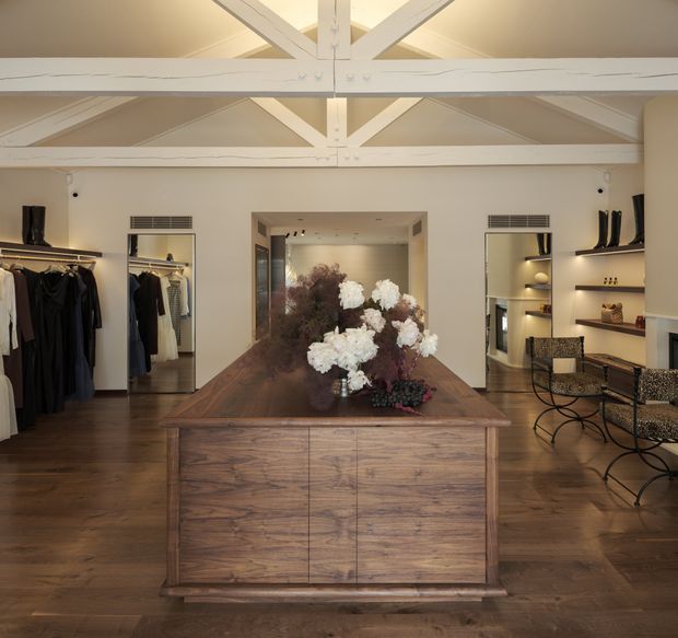 A refined boutique clothing showroom or dressing retail space with a large central timber display island, dark timber flooring, open clothing racks, illuminated shelving, and patterned lounge chairs. The room has a warm neutral palette and a prominent white-painted exposed timber truss ceiling with clerestory-like upper glazing.