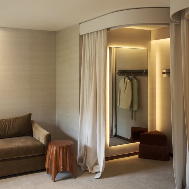 A softly lit, refined dressing area or walk-in wardrobe with full-height cream curtains framing an open alcove. A large illuminated mirror reflects hanging garments and built-in wardrobe storage, while a compact upholstered stool and draped side table sit on plush neutral carpeting. The room uses warm ambient lighting, textured pale wall finishes, and a restrained luxury aesthetic.
