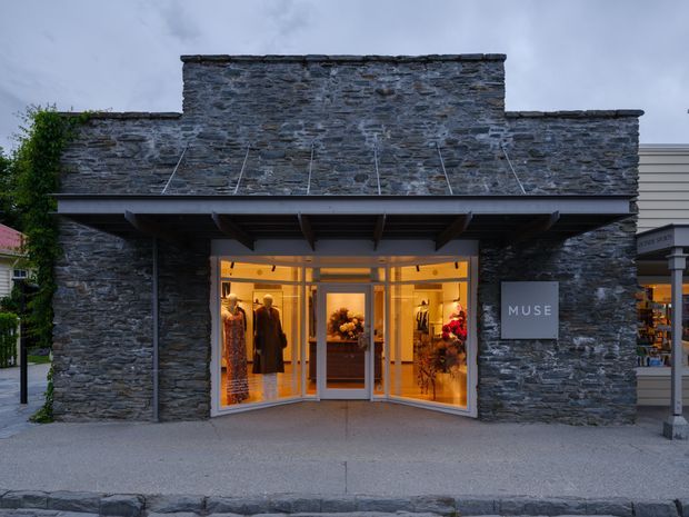 Street-facing commercial retail storefront with a symmetrical dark stone facade, stepped parapet-like massing, shallow metal canopy, and fully glazed entrance. Warm interior lighting reveals clothing displays and floral arrangements inside. The image is taken from the exterior on a paved pedestrian frontage.
