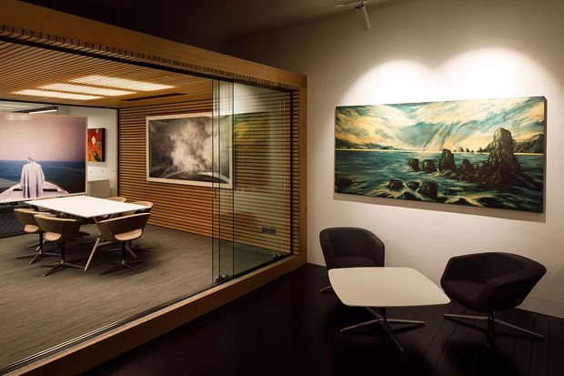 A refined contemporary office interior featuring a dark reception or lounge area in the foreground with a small white table and two dark upholstered armchairs. Through a full-height glazed partition with timber venetian-style screening, a conference room is visible with a white meeting table, multiple chairs, carpeted flooring, timber ceiling slats, and wall-mounted artwork. Warm integrated lighting and dark timber finishes create a polished corporate atmosphere.
