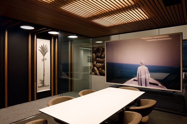A sophisticated contemporary dining or meeting room with a long white table surrounded by upholstered chairs. The space features dark wall panels with warm timber trims, a dramatic slatted timber ceiling with integrated lighting, glass partitions, carpeted flooring, and a large ocean-themed artwork. The overall atmosphere is intimate, refined, and moody.