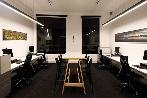 A clean, symmetrical modern office interior with two rows of workstations along the side walls, black ergonomic office chairs, white desks and cabinetry, large dark-framed windows, linear suspended lighting, and a central communal table with stools. White walls display several artworks, while dark carpet provides contrast.
