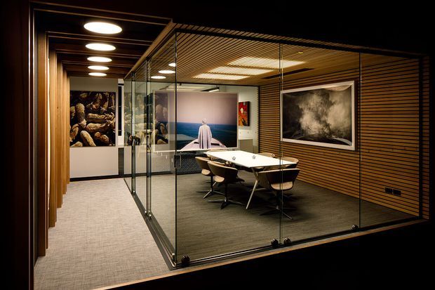 A sophisticated contemporary meeting room enclosed by clear glass partitions, with a long white conference table and dark swivel chairs. The room features warm timber slatted wall and ceiling finishes, integrated linear lighting, dark carpet, framed artwork, and a softly lit circulation area outside the glazed enclosure.
