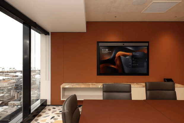 A contemporary conference or meeting room with floor-to-ceiling corner glazing overlooking an urban setting. The room features a large dark wood meeting table, leather chairs, warm terracotta wall panelling, a wall-mounted display, a pale stone or engineered-stone credenza, patterned flooring, and a suspended acoustic ceiling with recessed services.