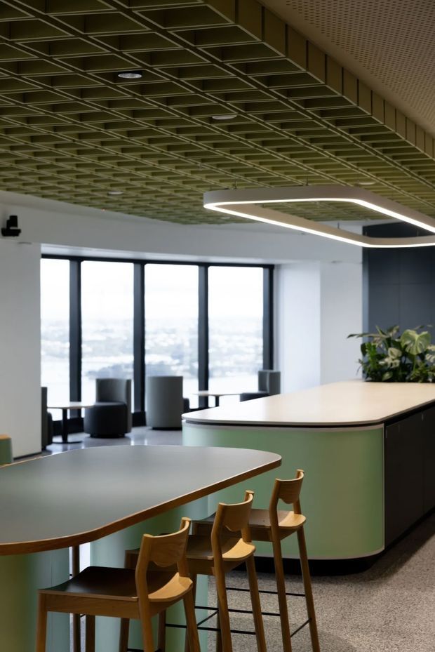 A contemporary commercial interior, likely a workplace café or reception breakout area, with curved pale-green counters, dark cabinetry, timber bar stools, lounge seating, expansive dark-framed windows, and a striking suspended geometric light. The ceiling features an exposed modular grid with recessed lighting, while terrazzo-like flooring and a planted counter create a refined, modern atmosphere.