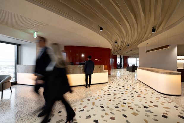 A spacious contemporary commercial lobby or reception area with a sculptural undulating timber ceiling, polished terrazzo floor with multicoloured stone aggregate, curved illuminated reception desks, and a glossy deep-red cylindrical feature wall. Large windows bring in daylight and reveal a distant cityscape. The space has refined hospitality-style detailing and active pedestrian movement.