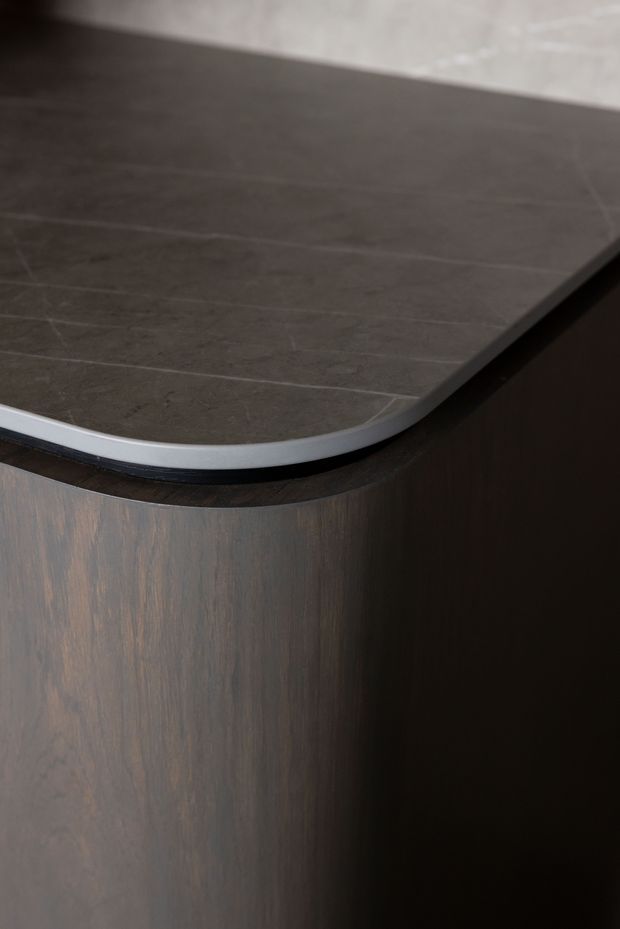 Close-up detail of a dark, rounded-edge countertop or benchtop with subtle stone-like veining, set over dark wood-grain cabinetry. The image focuses on the junction between the benchtop and curved built-in joinery.