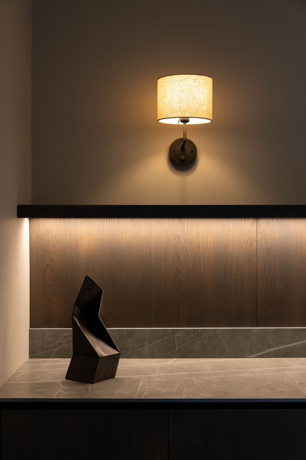A moody, refined interior vignette featuring a warm fabric wall sconce above dark timber wall panelling, a slim black ledge, and a stone display shelf with a sculptural dark abstract object. The lower section contains dark built-in cabinetry, while grey-brown veined stone forms the shelf and lower wall band.