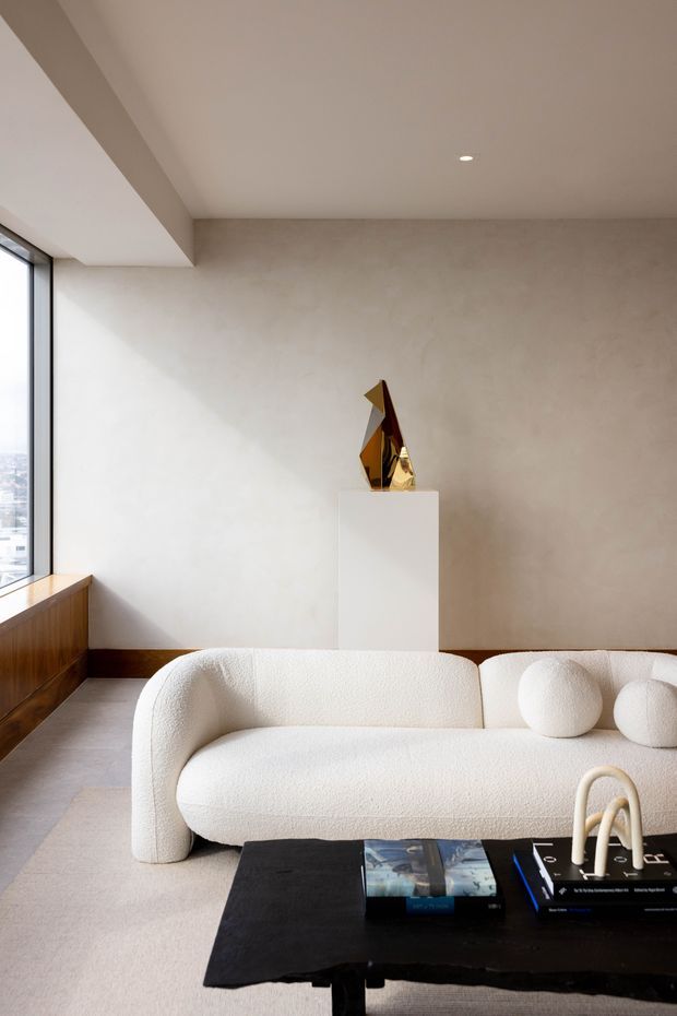 A minimalist living room with a sculptural cream boucle sofa, round cushions, a black coffee table with books, and a geometric metallic artwork displayed on a white plinth. The space has warm pale plaster walls, a light floor, extensive black-framed glazing, and restrained contemporary detailing.