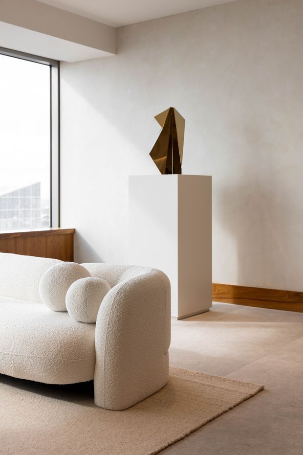 A softly lit contemporary living room featuring a sculptural cream boucle sofa with rounded arms, a neutral woven rug, pale tiled flooring, warm timber skirting and cabinetry, and a white pedestal displaying an angular metallic artwork. Large black-framed glazing brings in abundant daylight.