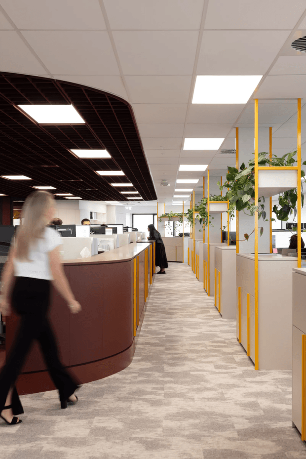A bright, contemporary open-plan office interior with rows of workstations and partitions, a curved dark burgundy reception or collaborative counter in the foreground, yellow metal-framed shelving and planter screens, abundant indoor greenery, patterned carpet, and a suspended grid ceiling with rectangular fluorescent light panels. People are present but blurred and secondary to the workspace.