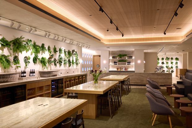 A sophisticated hospitality interior featuring a long bar and multiple marble-topped islands with timber cabinetry, extensive backlit shelving, upholstered dining chairs, and abundant vertical greenery. The room has a warm timber slatted ceiling with recessed cove lighting, black track lighting, polished stone surfaces, and a refined lounge-bar atmosphere.
