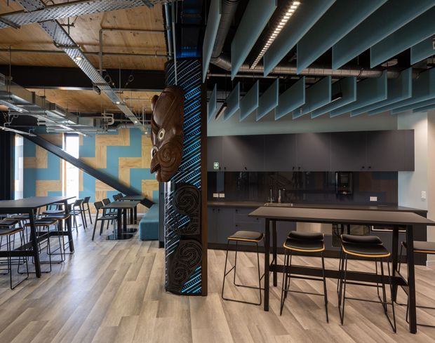 An open-plan contemporary commercial interior with a café or breakout kitchen area. The space features dark built-in cabinetry and a long island bench with bar stools, communal tables and chairs, exposed ceiling services, acoustic ceiling baffles, timber-look flooring, and a sculptural carved timber column element. Bright blue and natural timber wall accents add visual interest.
