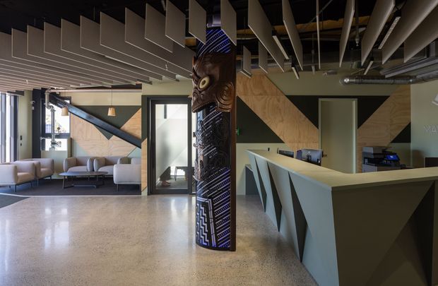 A contemporary commercial reception and lounge interior with a large angular reception desk, polished concrete or terrazzo-like flooring, geometric timber wall panels, exposed services, and dramatic suspended acoustic ceiling fins. A carved decorative column with Māori-inspired patterning forms a central feature, while a seating area and glazed entry are visible to the left.