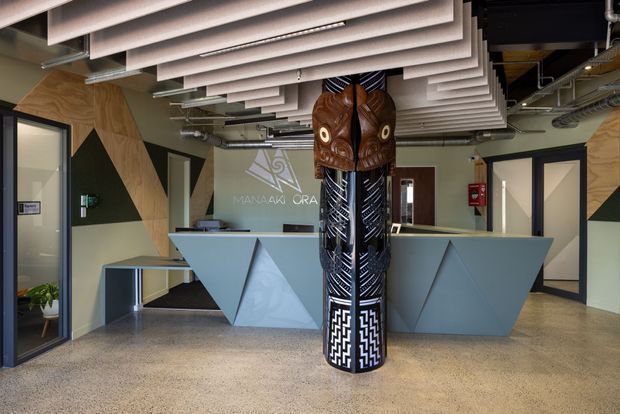 A contemporary commercial reception area with a sculptural geometric front desk, patterned cylindrical artwork or column, polished concrete or terrazzo-style flooring, timber and dark painted wall panels, and a ceiling fitted with suspended linear acoustic baffles. Glazed office doors and exposed services are visible around the lobby.