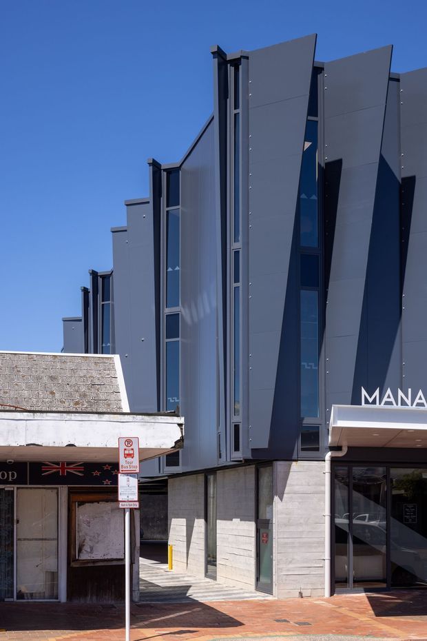 Street-level exterior view of a contemporary multi-storey commercial building with a highly articulated grey facade. Tall narrow glazing is set between angular projecting panels, with exposed concrete at the lower level and a glazed entrance canopy marked with partial signage. The building fronts a brick-paved urban footpath beside an older low-rise storefront under a clear blue sky.