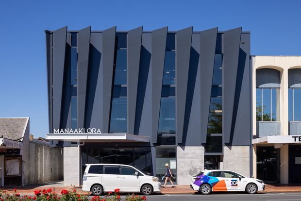 Street-level exterior view of a contemporary multi-storey commercial building with a dark grey facade articulated by repeated projecting angular fins, tall vertical glazing, and a projecting entrance canopy. Adjacent buildings and parked vehicles are visible along the streetscape.