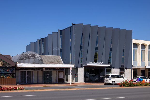 Street-level view of a contemporary multi-storey commercial building with a dramatic dark grey folded-panel facade, narrow vertical glazing, and a recessed ground-floor entrance. Adjacent low-rise commercial storefronts and an arched light-coloured building are visible along the streetscape, with a landscaped verge and clear blue sky.