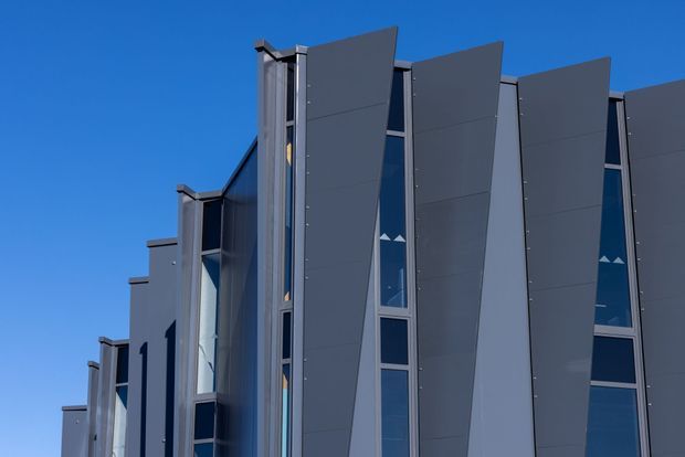 Close-up exterior view of a contemporary multi-storey building facade under a clear blue sky. The elevation features repeated tall, projecting dark grey vertical panels, narrow glazed openings, and silver-grey framing, creating a strong rhythmic and geometric composition.