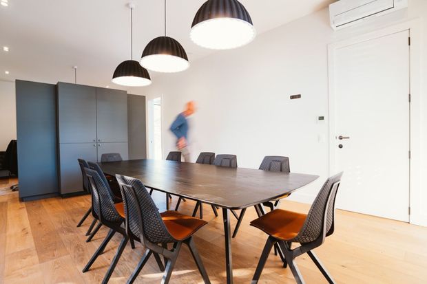 Bright, contemporary dining area with a long dark timber dining table surrounded by modern black-framed chairs with orange upholstered seats. The room has pale walls, light timber flooring, minimalist white cabinetry or storage along the left side, multiple black pendant lights, recessed ceiling lighting, and a white door with a wall-mounted split-system air conditioner above it.